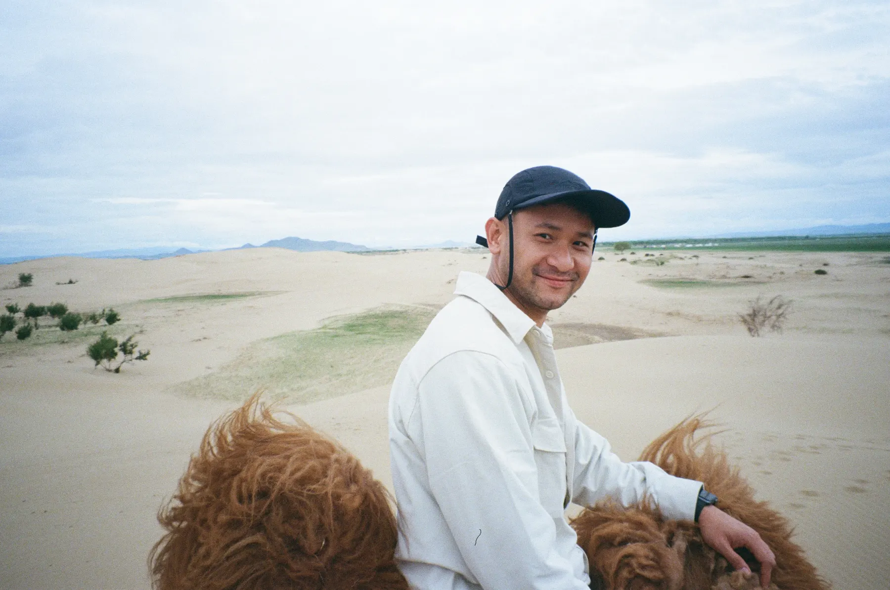 Adam in a cap and a light shirt, riding a Bactrian camel across sand dunes